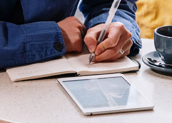 A man writing on a notebook while using his ipad