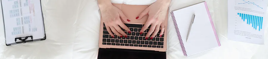 Person typing on a laptop while on bed with notebooks and papers